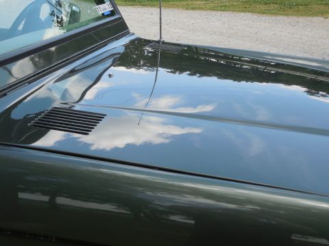 1975 Ford Bronco - Tight paint-quality shot down the passenger side of a dark green vintage Ford Bronco hood, showing sky and clouds mirrored in the glossy fi…