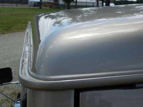 1975 Ford Bronco - Close-up looking across the front corner of a metallic tan/champagne hardtop roof, showing the glossy painted surface with tree reflections…