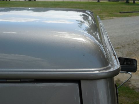 1975 Ford Bronco - Detail of the front corner of a metallic tan/champagne removable hardtop, showing sky and trees mirrored in the glossy paint, the formed dr…