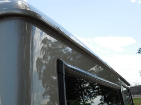 1975 Ford Bronco - Tight low-angle detail of the rear roof edge and drip rail on a light tan/beige hardtop, showing the glossy painted panel with trees reflec…