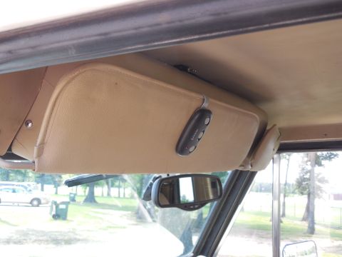 1975 Ford Bronco - Interior view looking up at the tan headliner and driver's sun visor of a classic SUV, with a three-button LiftMaster garage remote clipped…