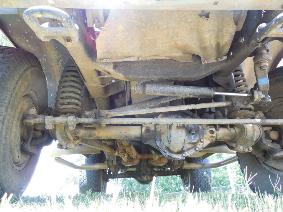 Underside of Ford Bronco front axle, leaf and coil springs, differential, and dusty rusty suspension over grass.