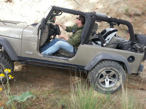 2005 Jeep Wrangler - A man drives a tan Jeep Wrangler on a dirt path with vegetation.