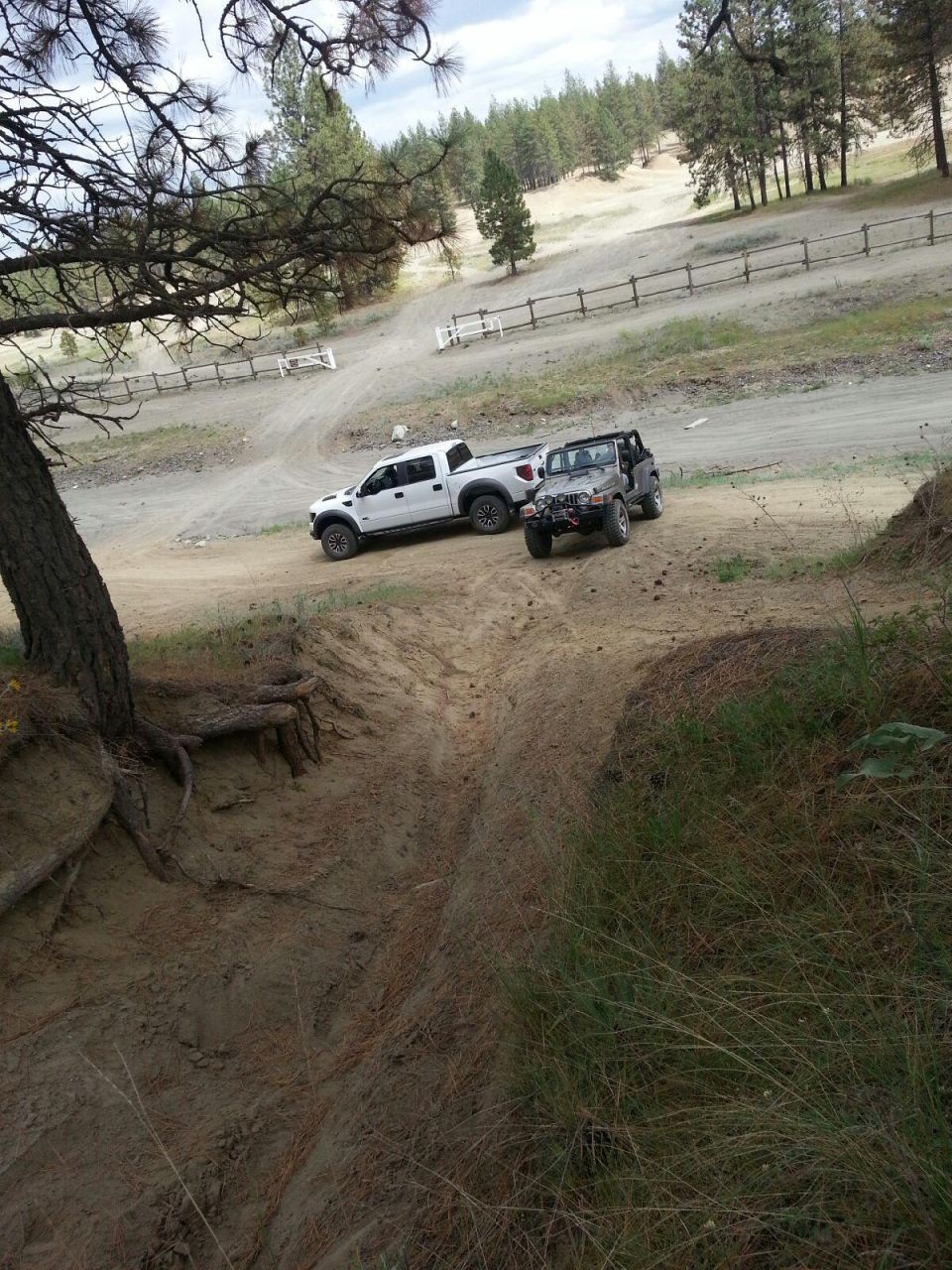 2005 Jeep Wrangler and a white pickup truck on a dirt trail with trees and hills in the background