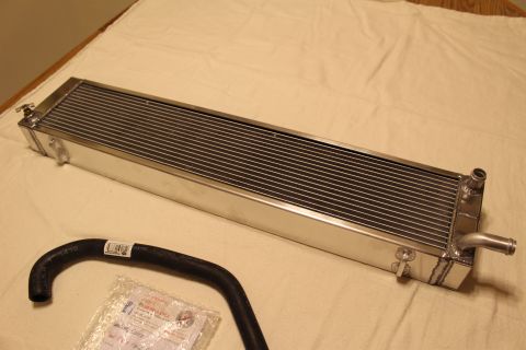 2003 Ford Crown Victoria - long polished aluminum heat exchanger lies on a tan cloth next to a black coolant hose and a bagged hardware kit.