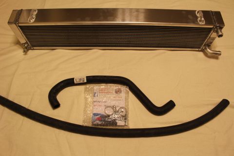 2003 Ford Crown Victoria - aluminum heat exchanger with mounting tabs lies above two black coolant hoses and a sealed hardware bag.