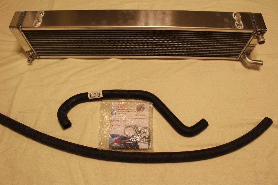 Aluminum heat exchanger with mounting tabs above two black hoses and a hardware bag
