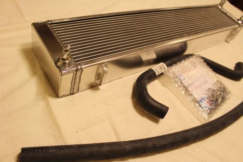 2003 Ford Crown Victoria - aluminum heat exchanger shown at an angle with a small brass fitting, beside a black hose and clear parts bag.