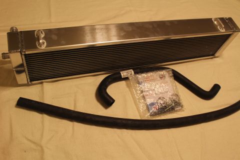 2003 Ford Crown Victoria - aluminum heat exchanger with mounting tabs lies above a curved black hose and a clear bag of clamps and parts.