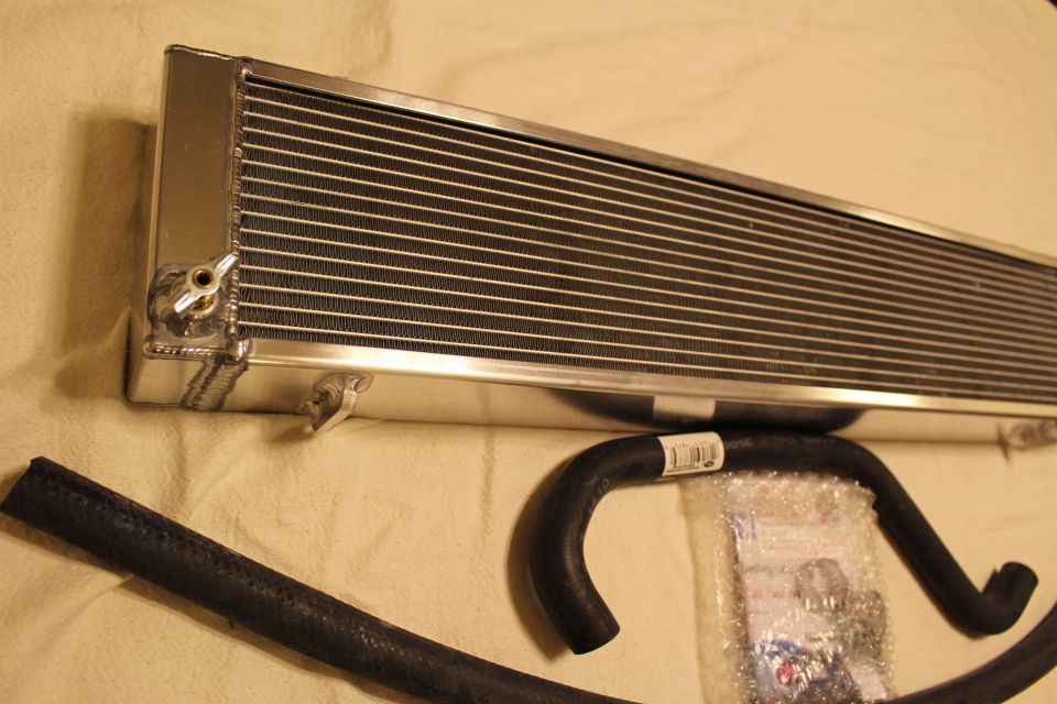 Angled aluminum heat exchanger with brass drain beside black hoses and a parts bag