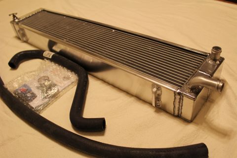 2003 Ford Crown Victoria - aluminum heat exchanger with twin barbed fittings sits at an angle beside a black hose and a sealed parts bag.