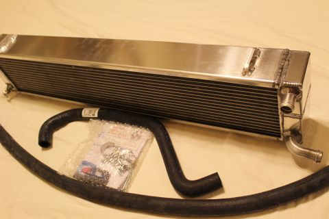 2003 Ford Crown Victoria - aluminum heat exchanger with twin barbed fittings sits at an angle beside a black hose and a sealed parts bag.