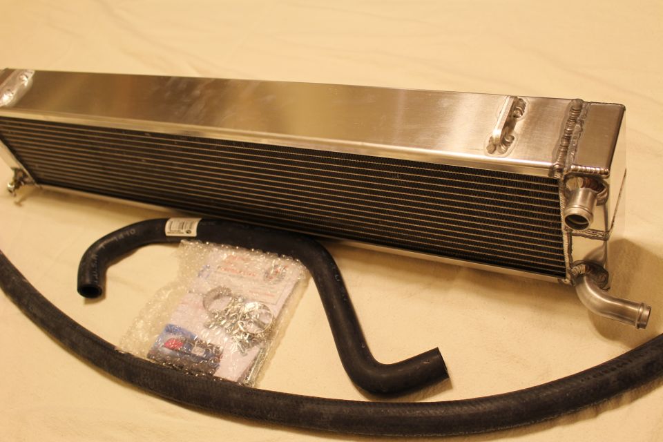 Angled aluminum heat exchanger with twin barbs beside a black hose and parts bag
