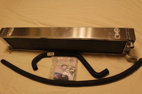 2003 Ford Crown Victoria - long polished aluminum heat exchanger lies flat beside a curved black rubber hose and a bag of clamps and hardware.