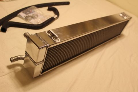 2003 Ford Crown Victoria - Lengthwise view of a polished aluminum heat exchanger with a corner barb fitting, a black hose and a bag of hardware nearby.