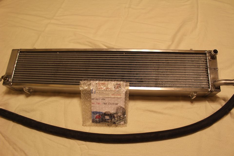 Polished aluminum heat exchanger with black hose and bagged hardware on tan sheet