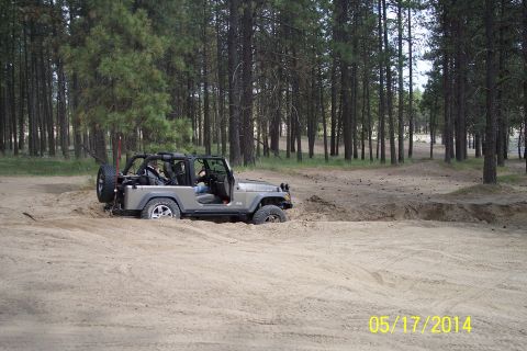 1990 Ford Bronco - A silver Jeep Wrangler navigates a sandy terrain amidst a forest, with its tires partially submerged in the sand.