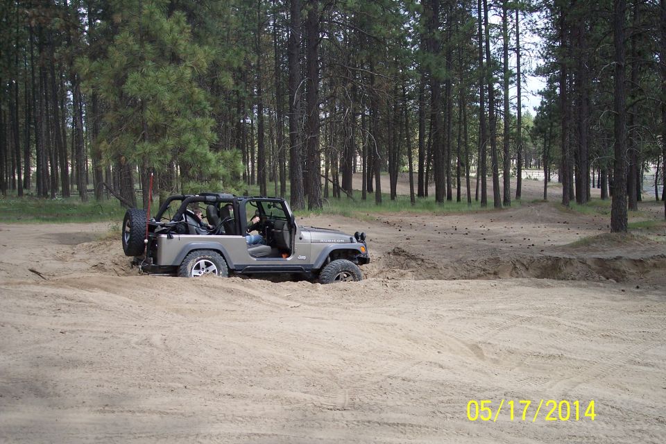 Silver Jeep Wrangler stuck in sand with spare tire on rear door, surrounded by pine trees