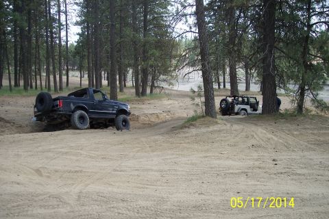1990 Ford Bronco - Two off-road vehicles navigate a sandy trail surrounded by pine trees.