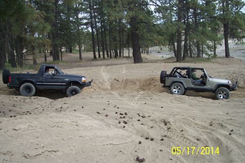 1990 Ford Bronco - Two off-road vehicles navigate a sandy terrain with trees in the background.