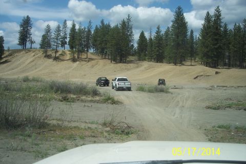 1990 Ford Bronco - Several vehicles navigate a dusty off-road trail surrounded by trees and sandy hills under a partly cloudy sky.