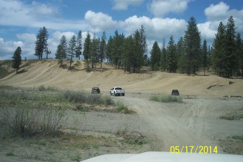 1990 Ford Bronco - Multiple vehicles navigate a sandy terrain with sparse vegetation and a backdrop of pine trees under a cloudy sky.