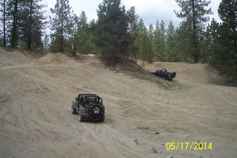 1990 Ford Bronco - Two off-road vehicles navigate a sandy terrain with trees in the background.
