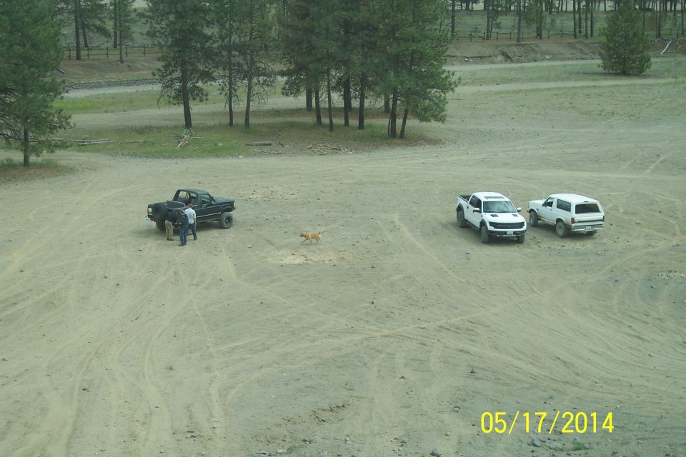 Black pickup truck with people nearby, white pickup truck, and white SUV parked in a dirt lot with trees in background