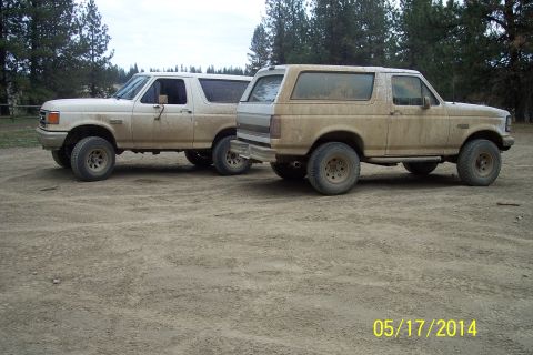 1990 Ford Bronco - Two dusty Ford Bronco SUVs are parked side-by-side on a dirt lot with trees in the background.