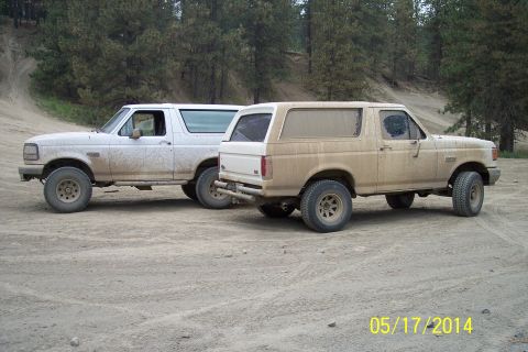 1990 Ford Bronco - Two Ford Broncos, one white and one tan, are parked side-by-side on a dirt road with trees in the background.
