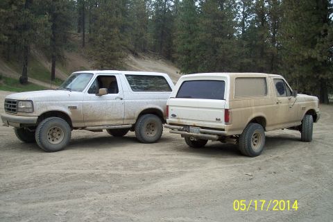 1990 Ford Bronco - Two Ford Broncos are parked on a dirt road, with trees in the background. The vehicle on the left is facing forward, while the one on the r…