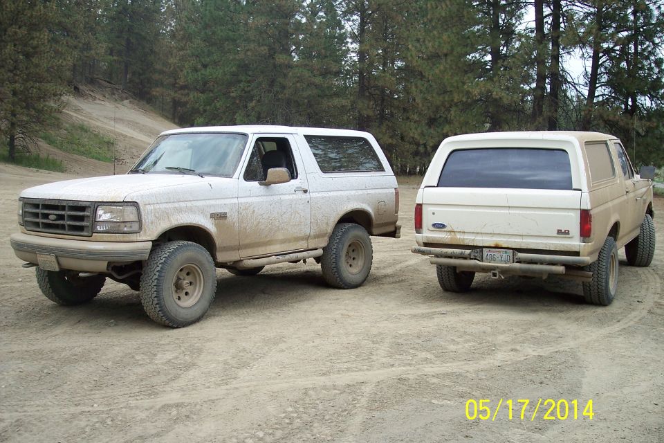 Two white Ford Broncos parked on a dirt road, one facing front-left, the other facing rear-right. Both appear to be from the early 1990s