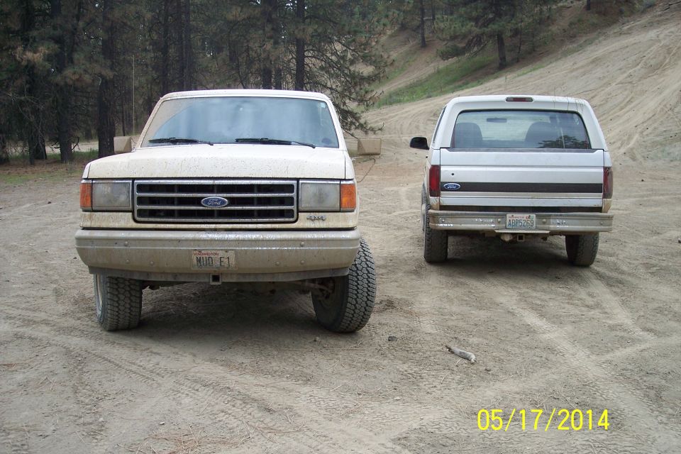 Two Ford Broncos parked on a dirt road, one facing front, the other facing rear. The front Bronco is a 1987 model