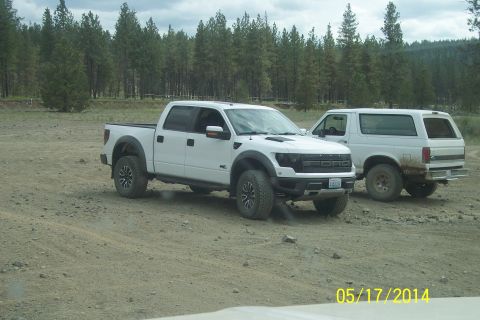 1990 Ford Bronco - Two white Ford vehicles, a Raptor pickup and a Bronco SUV, are parked on a dirt road with trees in the background.