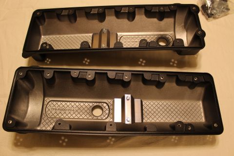 2003 Ford Crown Victoria - pair of black plastic valve covers lie interior-side-up, one cast 'TFS-FMOD-VC-L' with a polished metal clip across it.