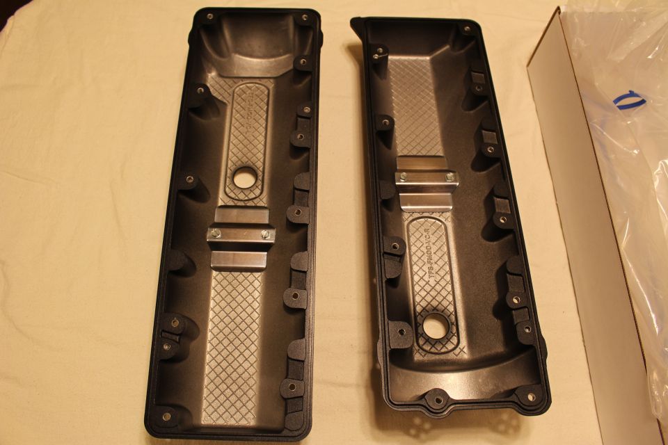 Two black valve covers placed side by side with undersides up near a cardboard box