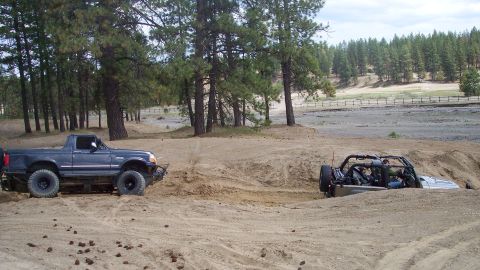 2005 Jeep Wrangler - Two off-road vehicles navigate a sandy terrain with trees and a fence in the background.