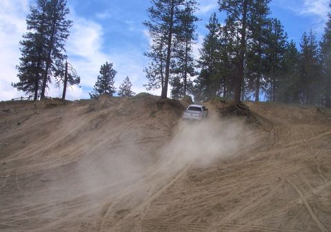 2005 Jeep Wrangler - A white pickup truck navigates a dusty, off-road trail surrounded by pine trees under a partly cloudy sky.