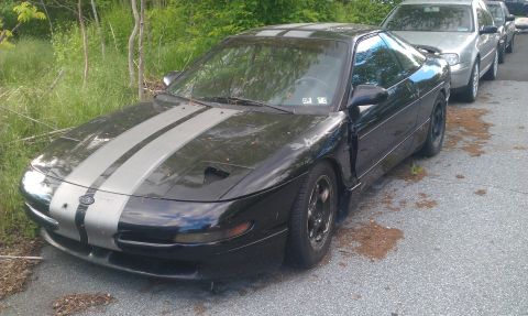 1993 Ford Probe - A black 1993 Ford Probe with racing stripes is parked on a paved surface next to other vehicles.