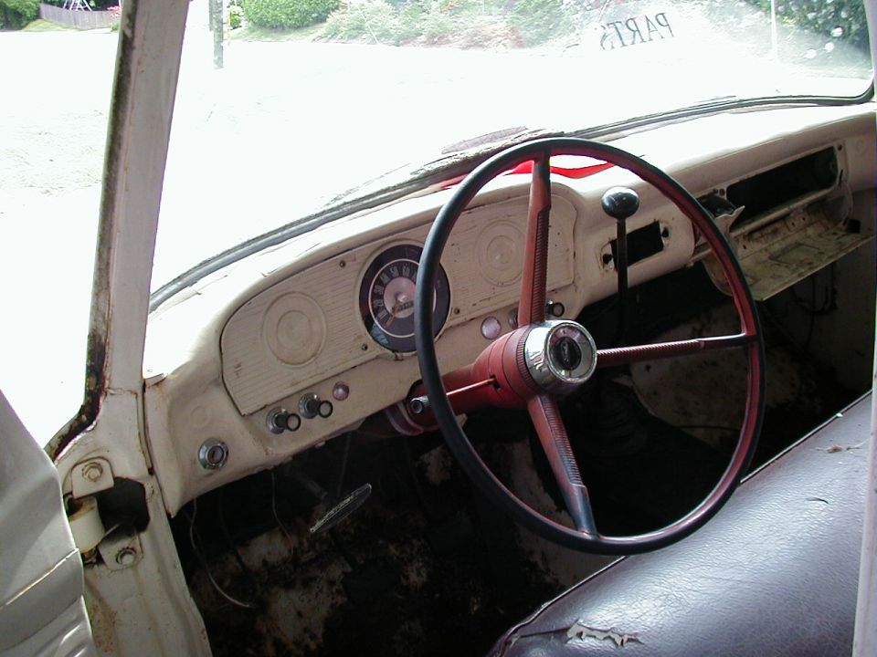 Vintage Ford truck interior with red steering wheel, cream dash, round gauge, and rusty bare floor; 'PARTS' lettering on windshield.