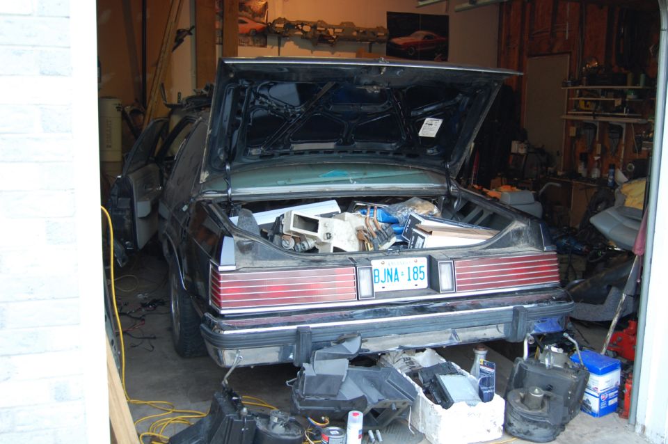 Rear view of a dark sedan in a garage, trunk open and loaded with car parts. License plate BJNA-185