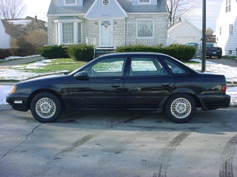 1973 Ford Ranchero - A black sedan is parked on a residential street with a house in the background. Another vehicle is visible further down the street.
