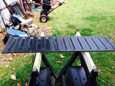 1979 Ford Bronco - A black tailgate is displayed outdoors on sawhorses, with a pile of scrap metal and a lawn spreader in the background.