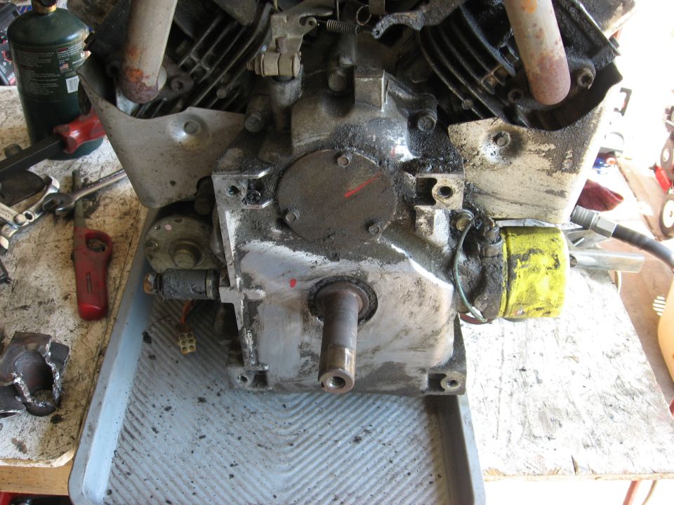 Engine block with oil residue, yellow filter, and metal shaft. Tools and a propane tank are visible in the background