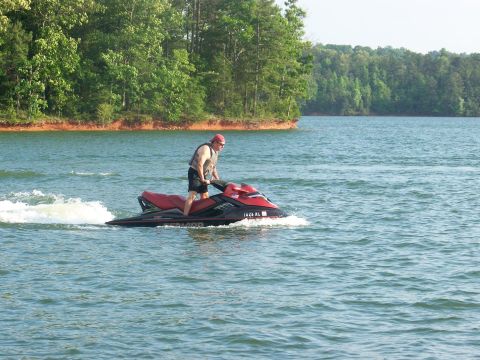 2006 Sea-Doo 230 Wake - A man rides a red and black jet ski on a lake with trees in the background.