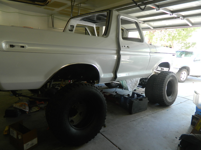 White 1978 Ford Bronco body with large tires, undergoing restoration in a garage. Another white SUV is visible in the background