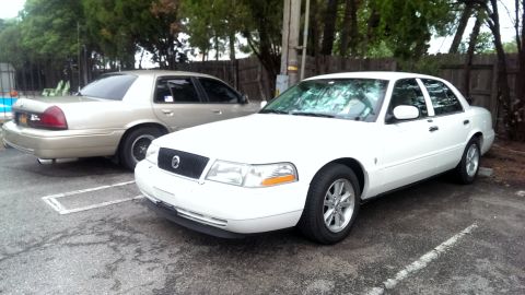2000 Mercury Grand Marquis - Two Mercury Grand Marquis sedans are parked outdoors, one white and one gold, with trees and a fence in the background.
