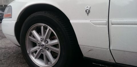 2000 Mercury Grand Marquis - Close-up of a white 2000 Mercury Grand Marquis showing the V8 emblem and a silver alloy wheel on the front fender.
