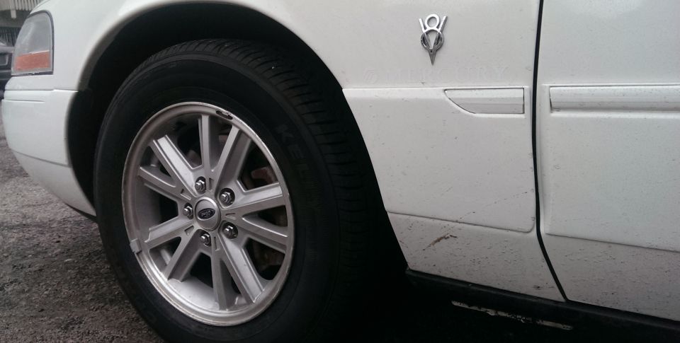 White car fender with a V8 badge, showing a silver multi-spoke wheel and tire