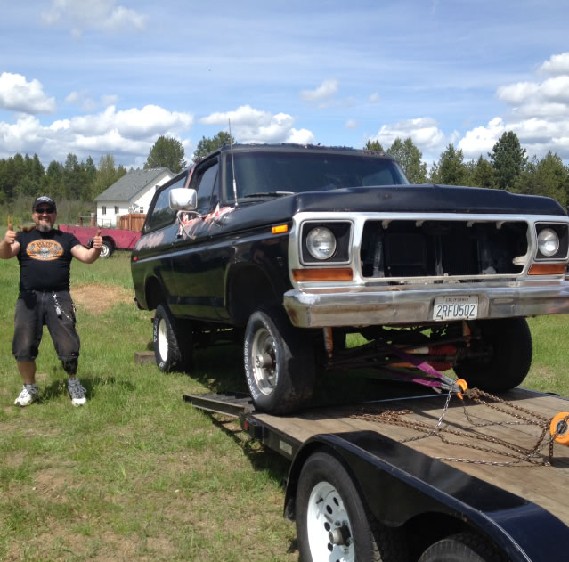 Guy giving thumbs up next to a black 1978 Ford Bronco on a trailer. Grille is missing